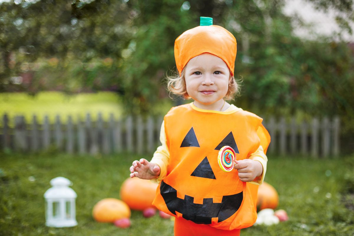Toddler wearing a pumpkin Halloween costume holding a lollipop outdoors.